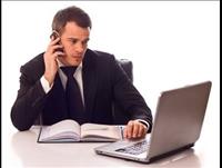 Man at his desk holding his phone to his ear while his other hand is typing on a laptop