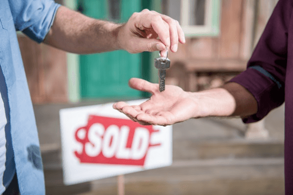 Hand passing over keys to someone else with a red "sold" sign in the background