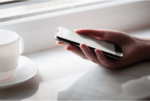 A hand holding a cell phone with a tea cup to the side